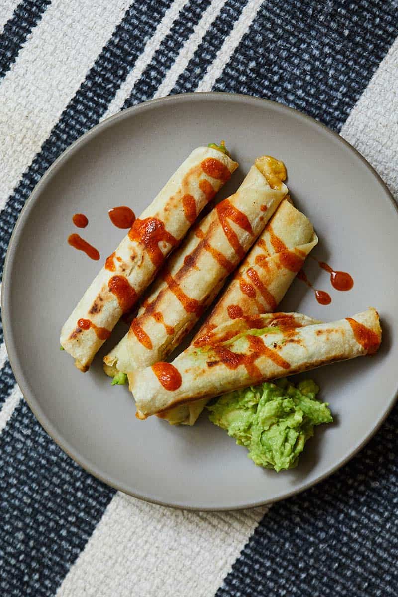 Avocado Hummus Taquitos garnished with hot sauce and side of guacamole on a gray plate