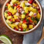 Pinterest graphic of a bowl of mango salsa with lime wedges and a wooden spoon surrounding it.