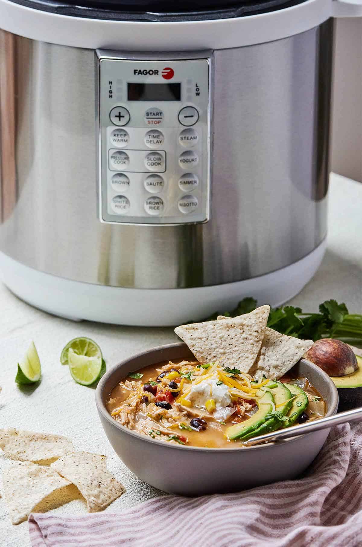 A bowl of chicken tortilla soup topped with chips and avocado in front of a pressure cooker.