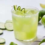 A glass of cucumber margarita with salt on the rim and a ribboned cucumber as garnish.