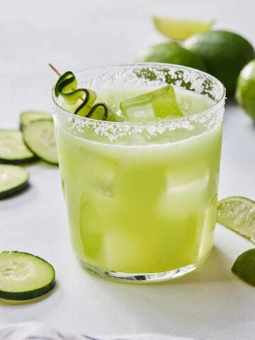 A glass of cucumber margarita with a salted rim and cucumbers slices around.