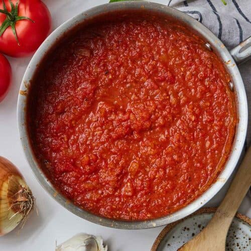 Overhead view of a saucepan of marinara sauce beside a wooden spoon, sprigs of basil, garlic cloves, and tomatoes.