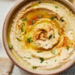 Pinterest graphic of an overhead view of a bowl of creamy hummus with paprika, olive oil, and garnish.
