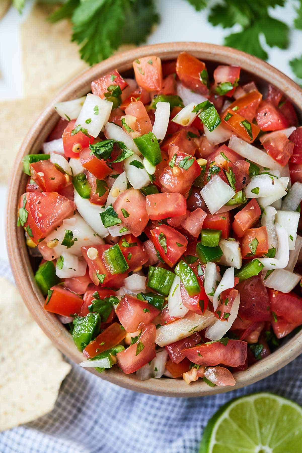 Overhead view of a bowl of pico de gallo beside chips, cilantro, and a lime.