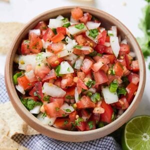 Overhead view of a bowl of pico de gallo in a brown bowl beside some chips and lime.