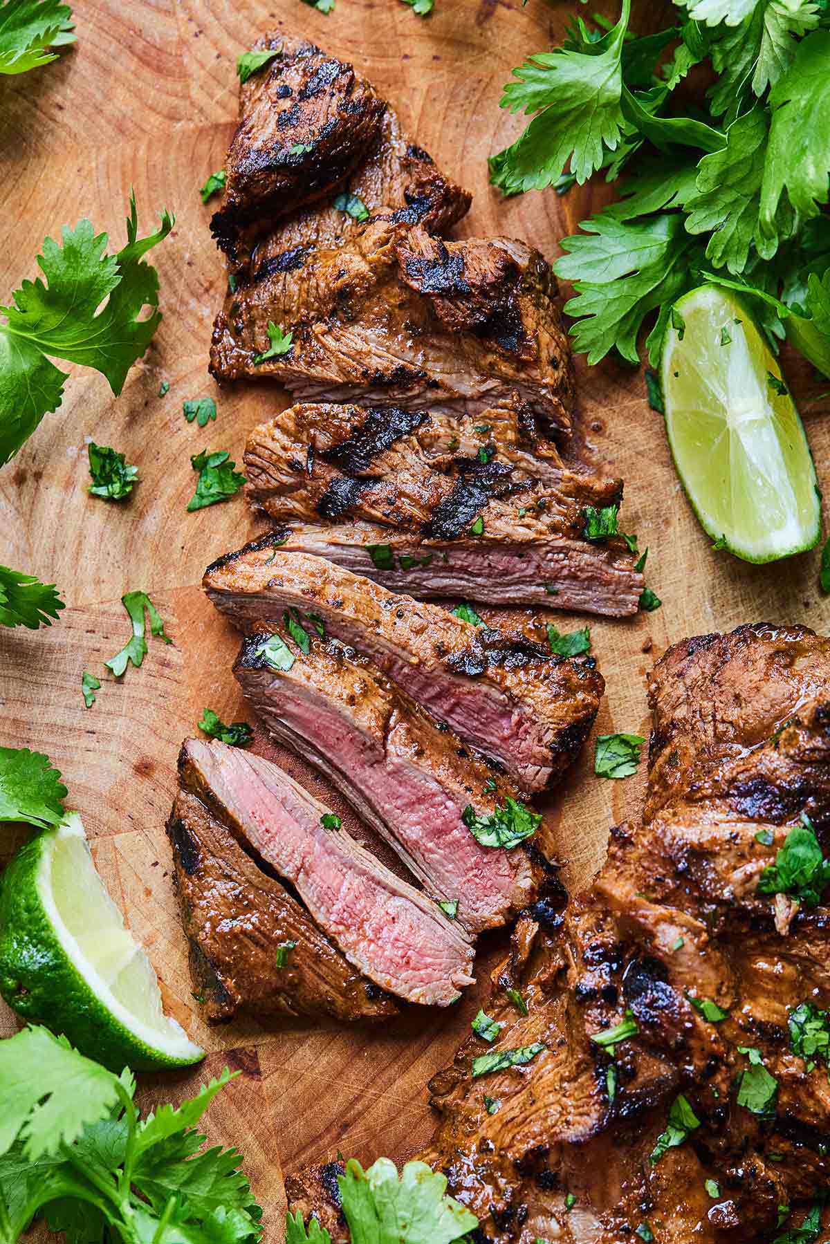 Overhead view of a sliced carne asada on a cutting board surrounded by cilantro leaves and lime wedges.