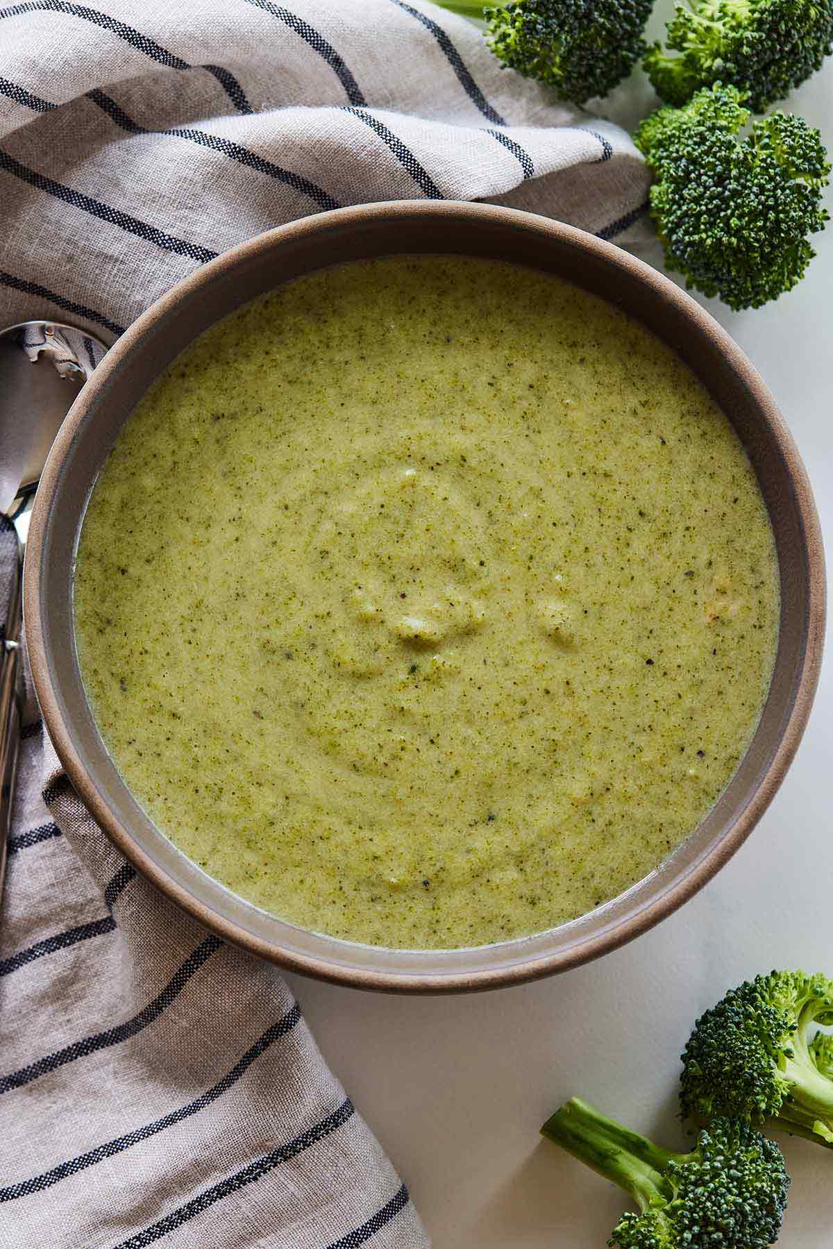 Overhead view of cream of broccoli soup beside a striped linen and broccoli florets.