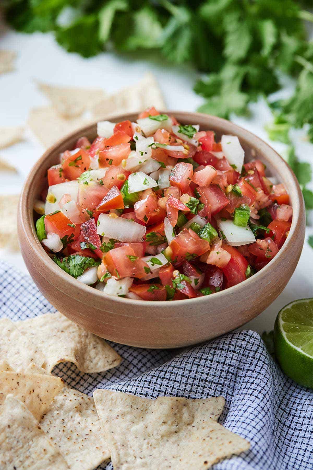 A bowl of pico de gallo by a linen napkin with tortilla chips on top and a cut lime.