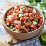 Pinterest image of a brown bowl of pico de gallo by a linen napkin with chips on top by a bunch of cilantro.