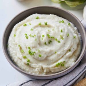 A grey bowl of mashed cauliflower with chives on top with a linen napkin beside it.