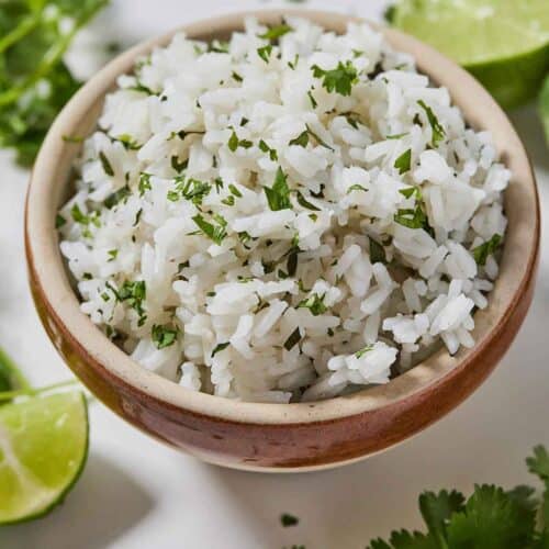 Overhead view of a bowl of cilantro lime rice with a couple of cut limes and fresh cilantros on the table.