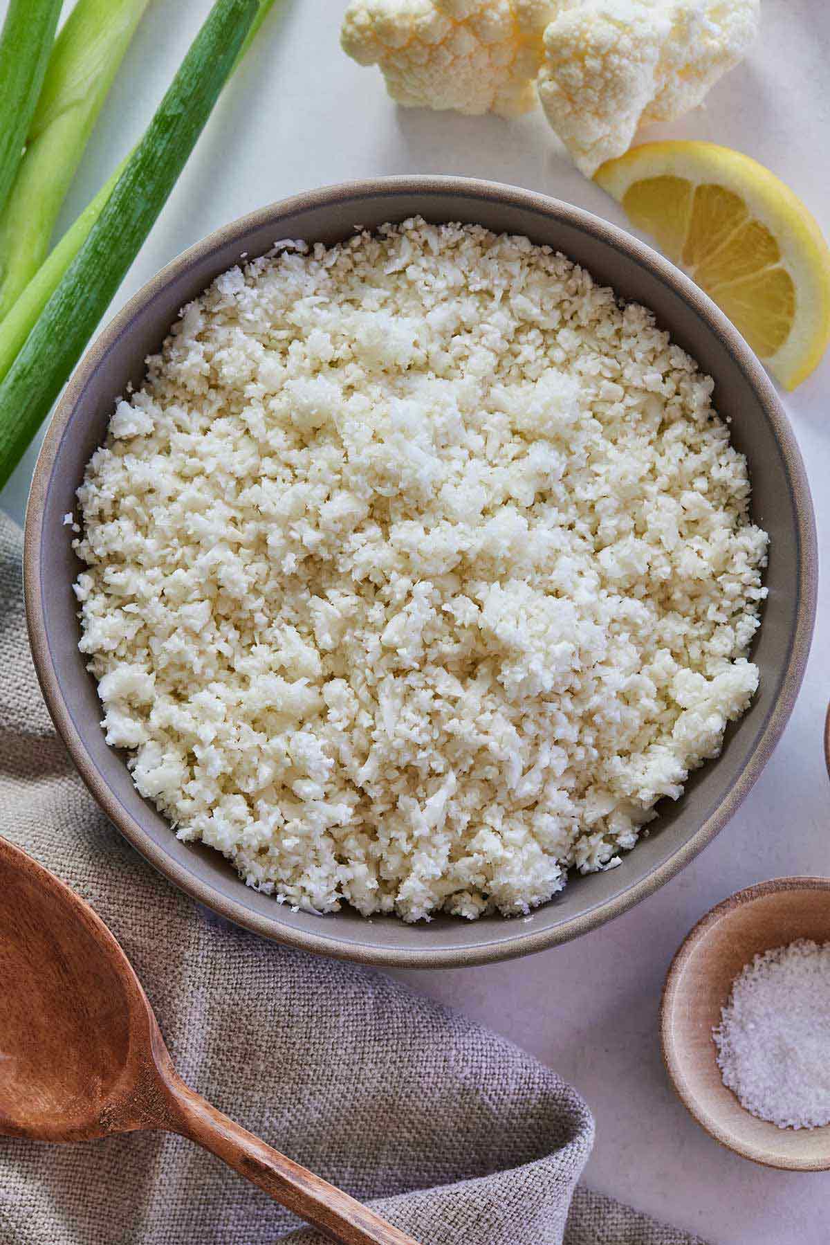 Overhead view of a bowl of cauliflower rice with green onions, a lemon wedge, and cauliflower florets around it with a pinch bowl of salt.