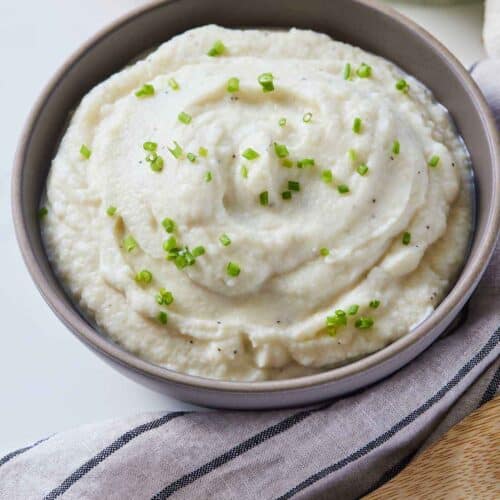 A bowl of mashed cauliflower with chives on top by a linen napkin and wooden spoon beside it.