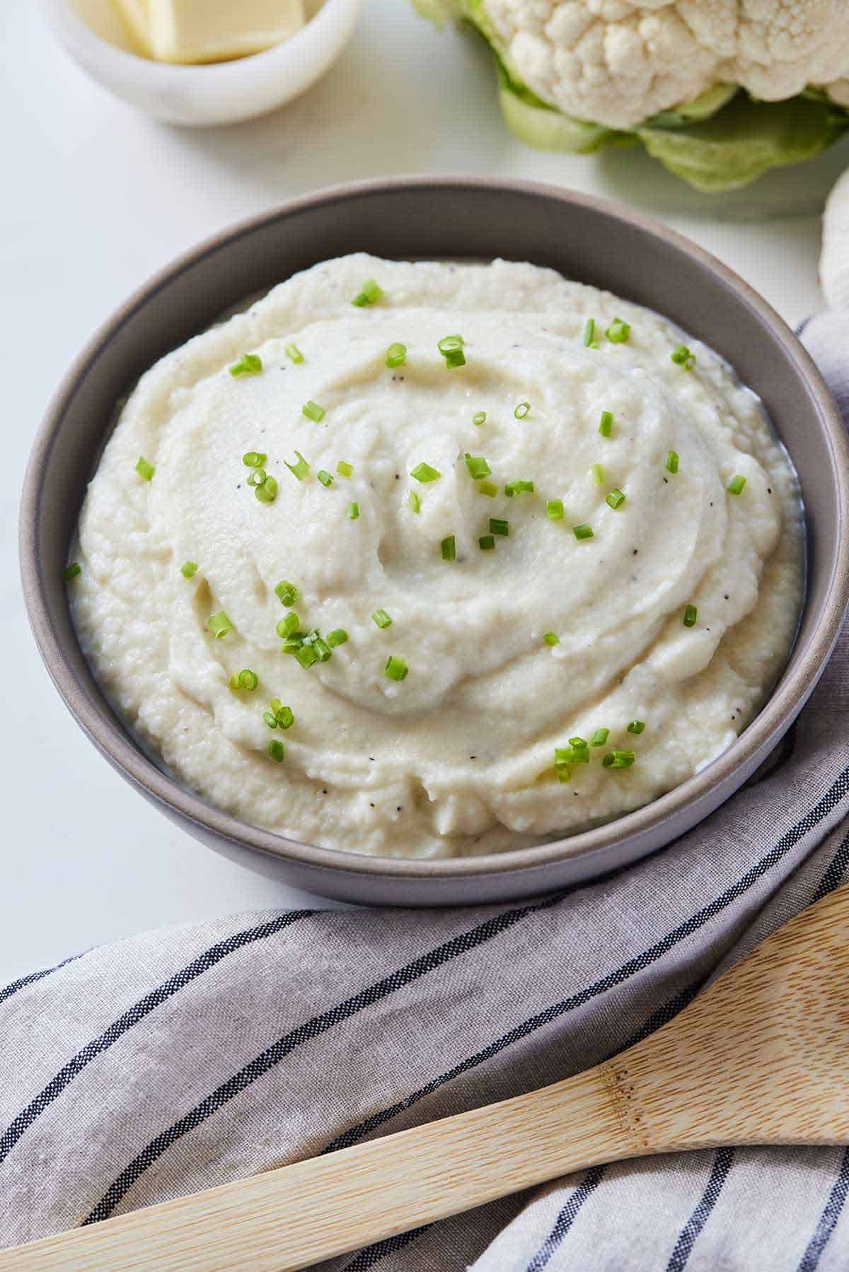 A bowl of mashed cauliflower with chives on top by a linen napkin and wooden spoon beside it.
