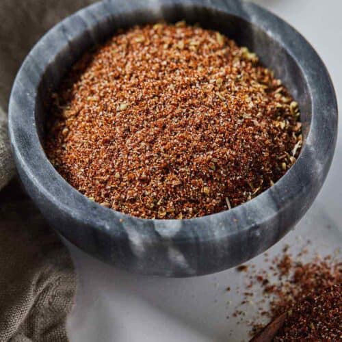 A grey pinch bowl of taco seasoning with a wooden spoon containing some beside it.