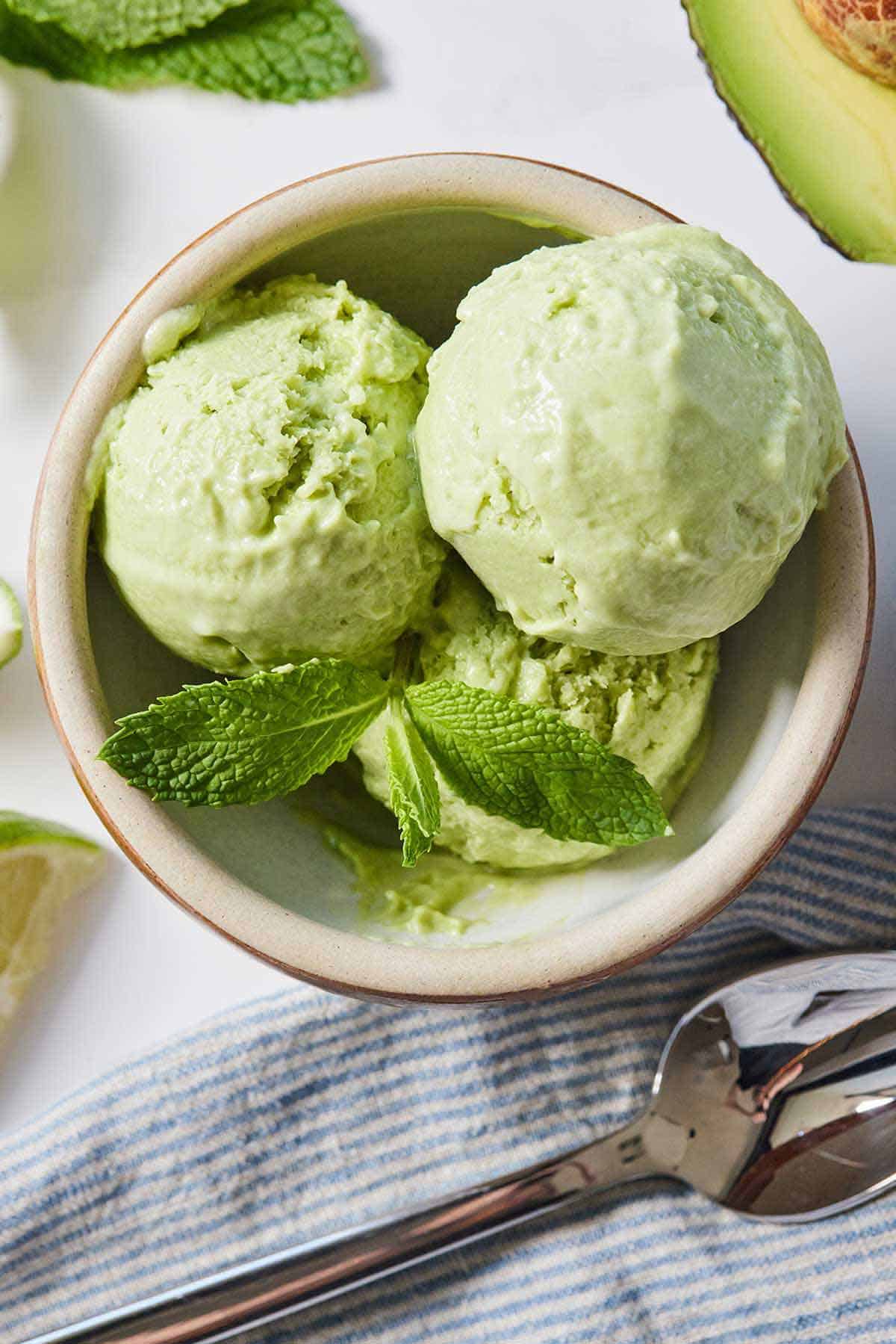 Overhead view of multiple scoops of avocado ice cream in a bowl with mint on top with a linen napkin and spoon beside it.