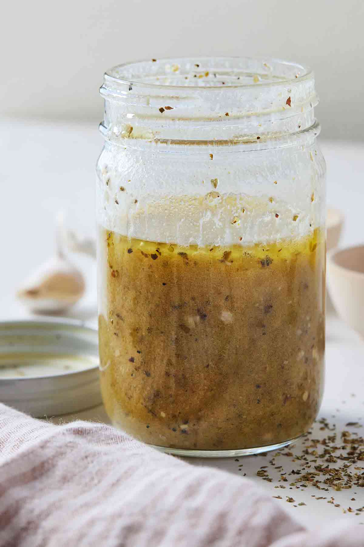 A mason jar of red wine vinaigrette with a striped linen napkin in front with some dried oregano scattered near the jar.