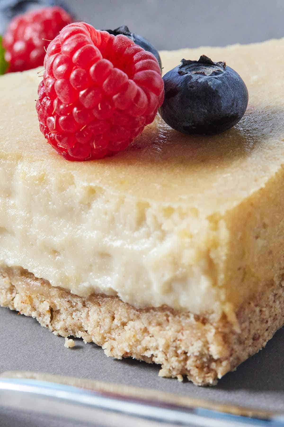 Close up photo of a slice of vegan cheesecake, showing the crust and filling, with a raspberry and blueberry on top.