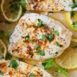 Close up of a piece of baked cod with fresh parsley and slices of lemon around it.