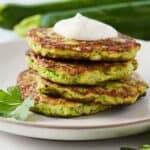 Side view of a stack of four zucchini fritters on a plate with parsley as garnish.