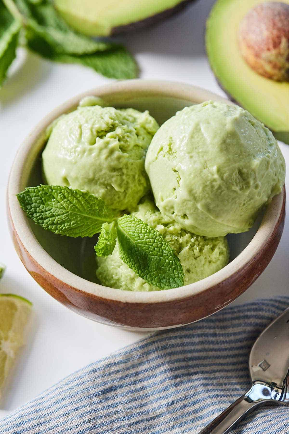 A bowl of avocado ice cream with fresh mint surrounded by cut avocado, lime wedges, and a linen napkin.