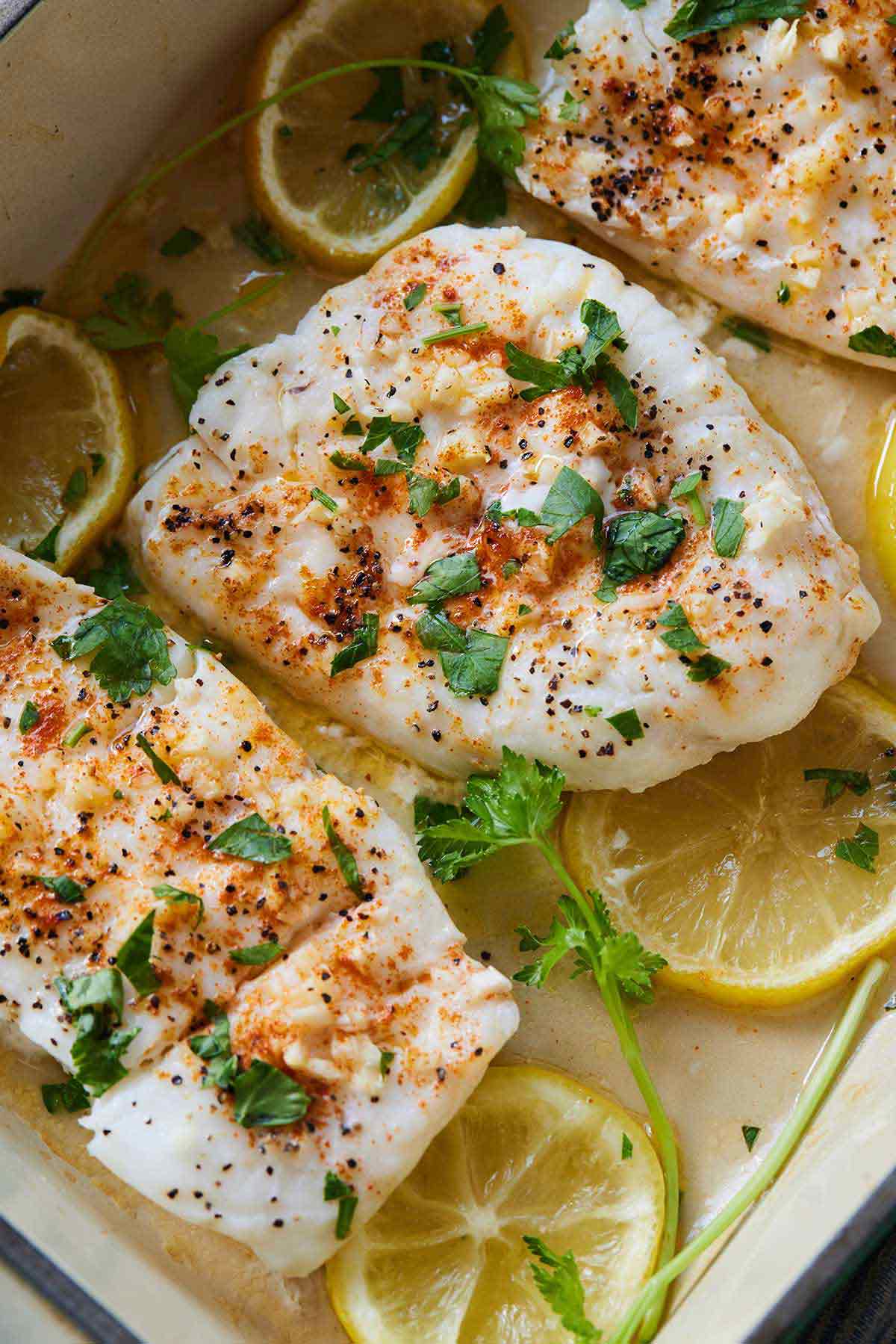 Overhead view of pieces of baked cod in a baking dish with fresh herbs and lemon slices.