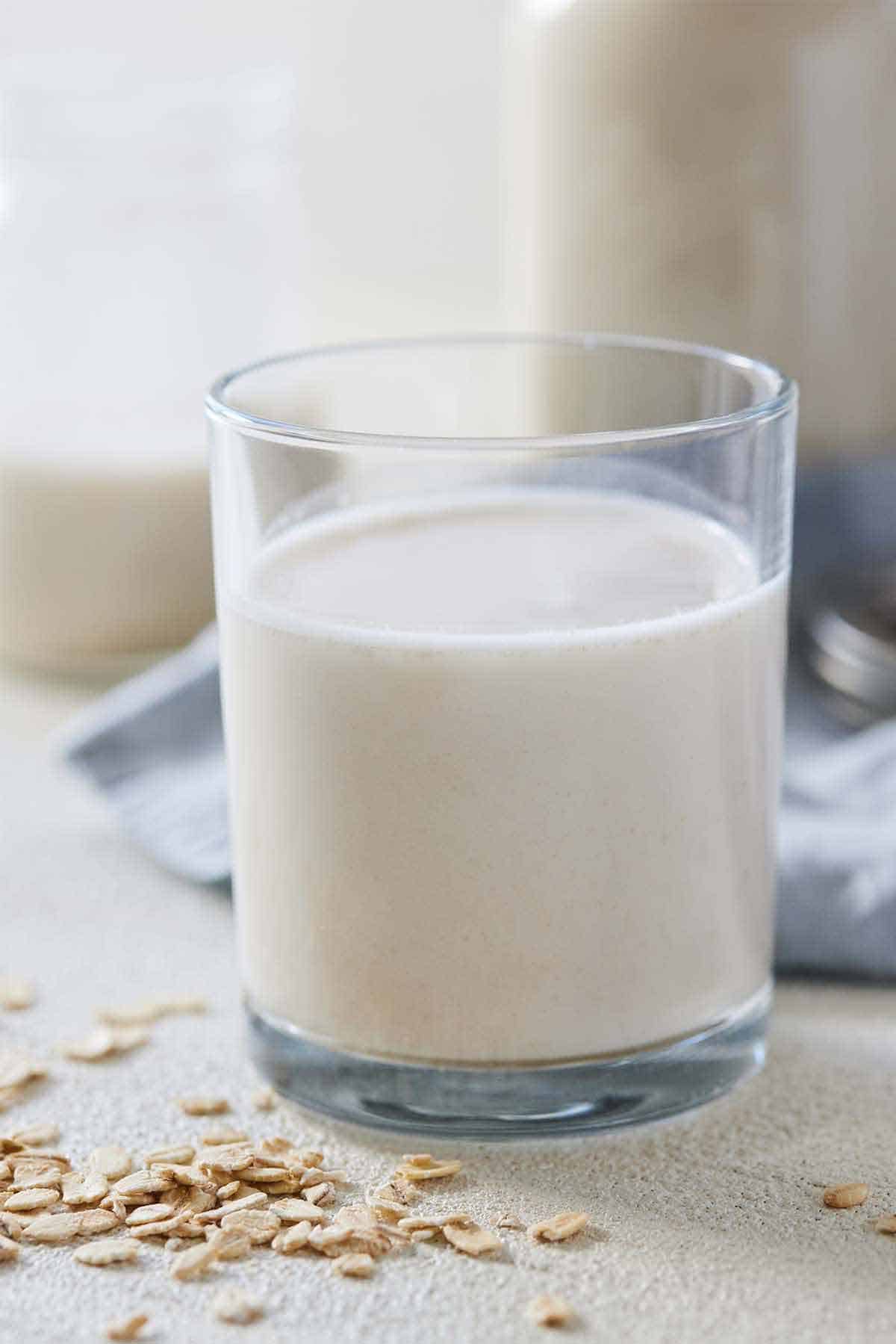A glass of oat milk with some oats scattered in front with a blue linen in the back.