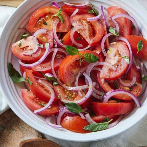 Overhead view of a bowl of tomato salad with wooden serving utensils beside it with some sliced bread nearby.