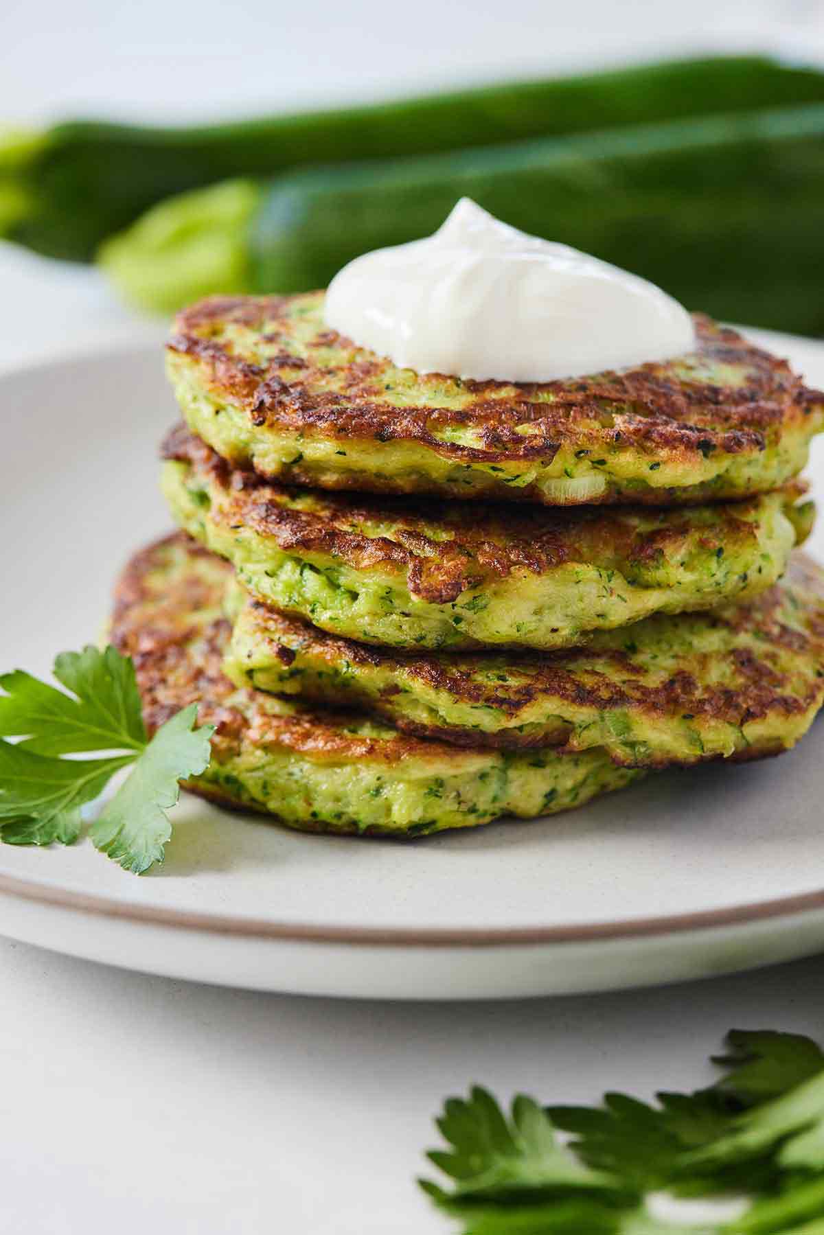 A plate of zucchini fritters with sour cream on top and some zucchinis in the background, out of focus.