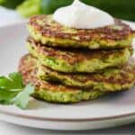 Pinterest graphic of a stack of four zucchini fritters with a dollop of sour cream on top.