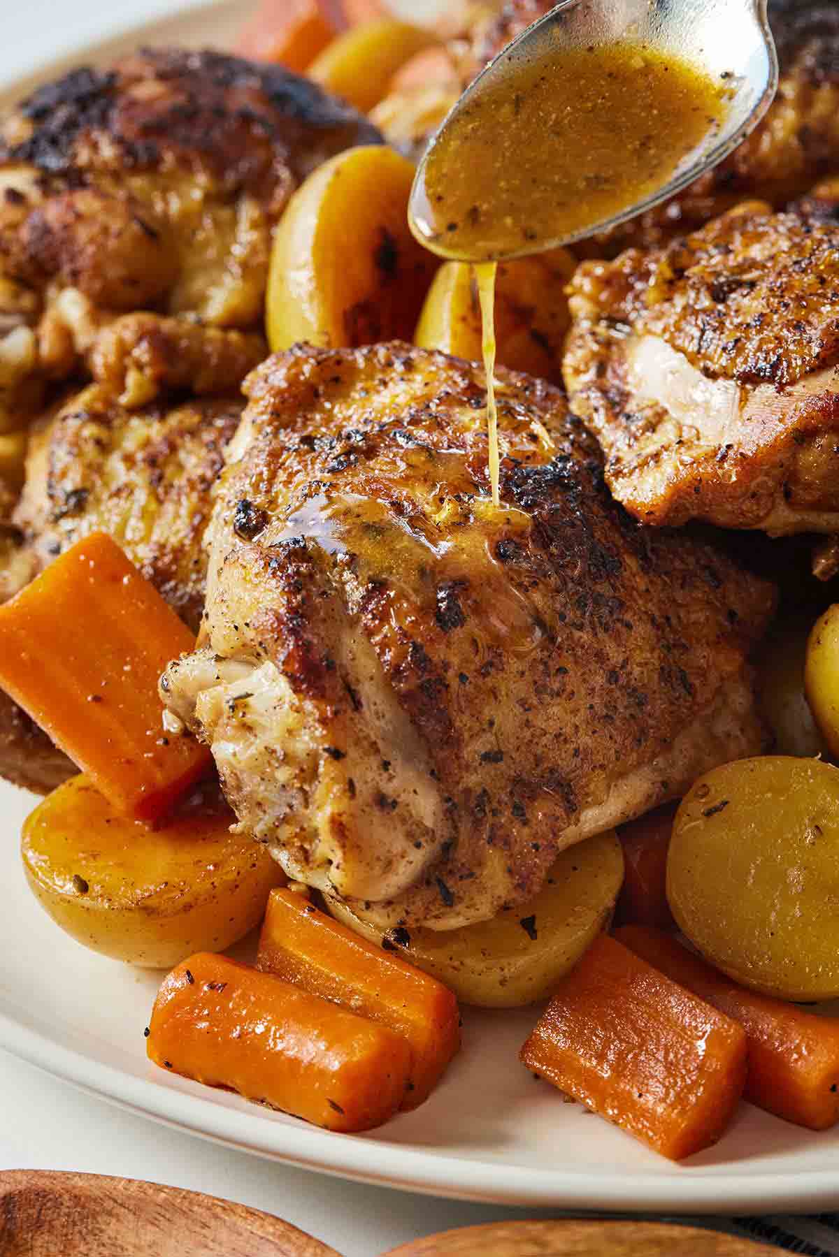 A spoon drizzling sauce over chicken thighs on a plate with carrots and potatoes.