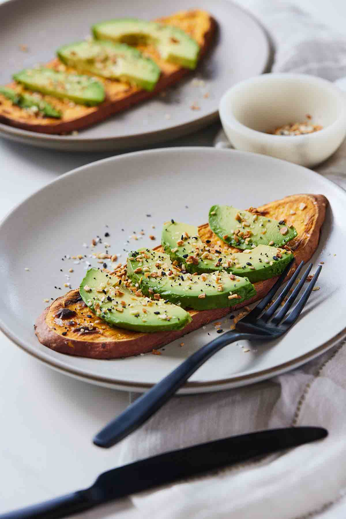 Two plates with one in focus with sweet potato toast and sliced avocado with a fork and knife.