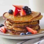 Close up of a stack of oatmeal banana pancakes with fruits on top and maple syrup.