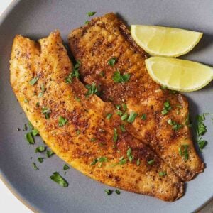 A plate with pan fried tilapia and two lemon wedges and herb garnish.