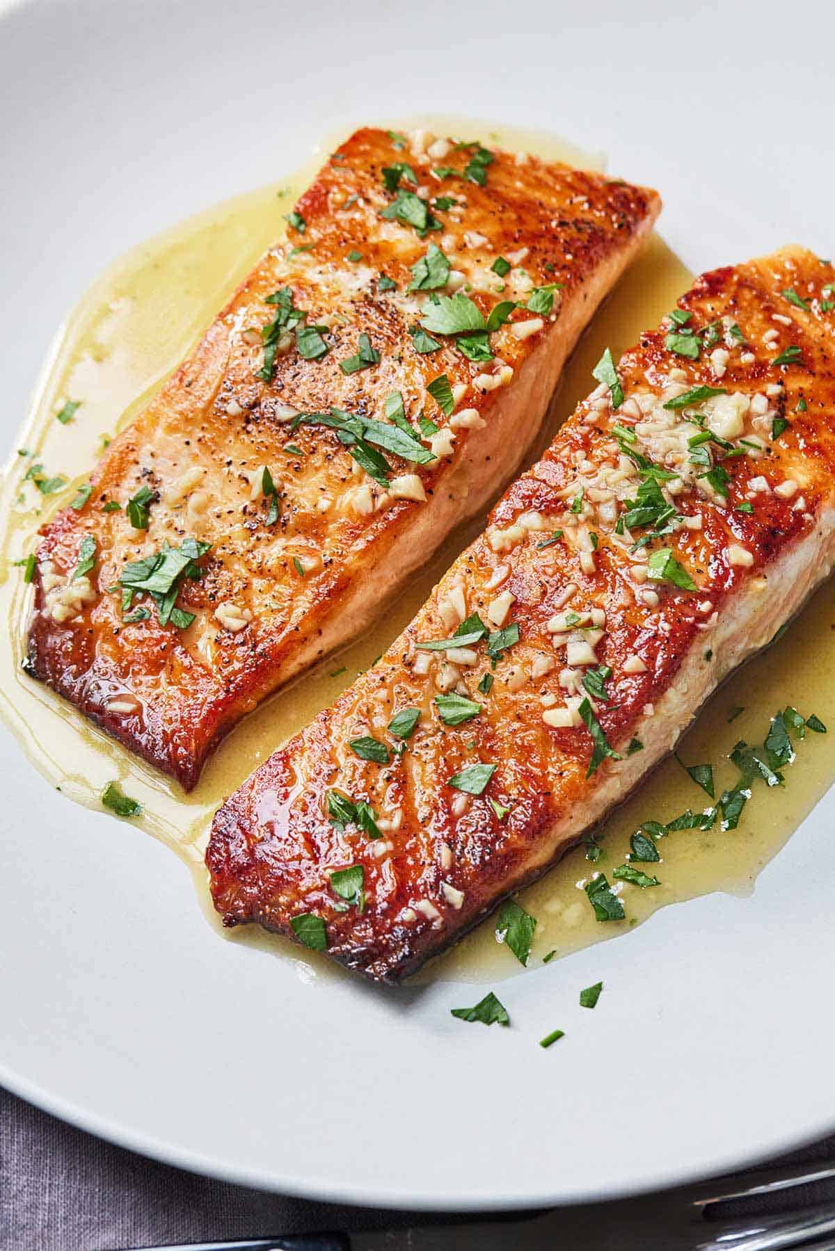 Two pan seared salmon fillets in a plate with a butter sauce poured over top of them. The top of the fillets have minced garlic and chopped parsley.