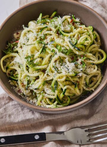A bowl of zucchini noodles with parmesan and basil on top with a fork beside it.