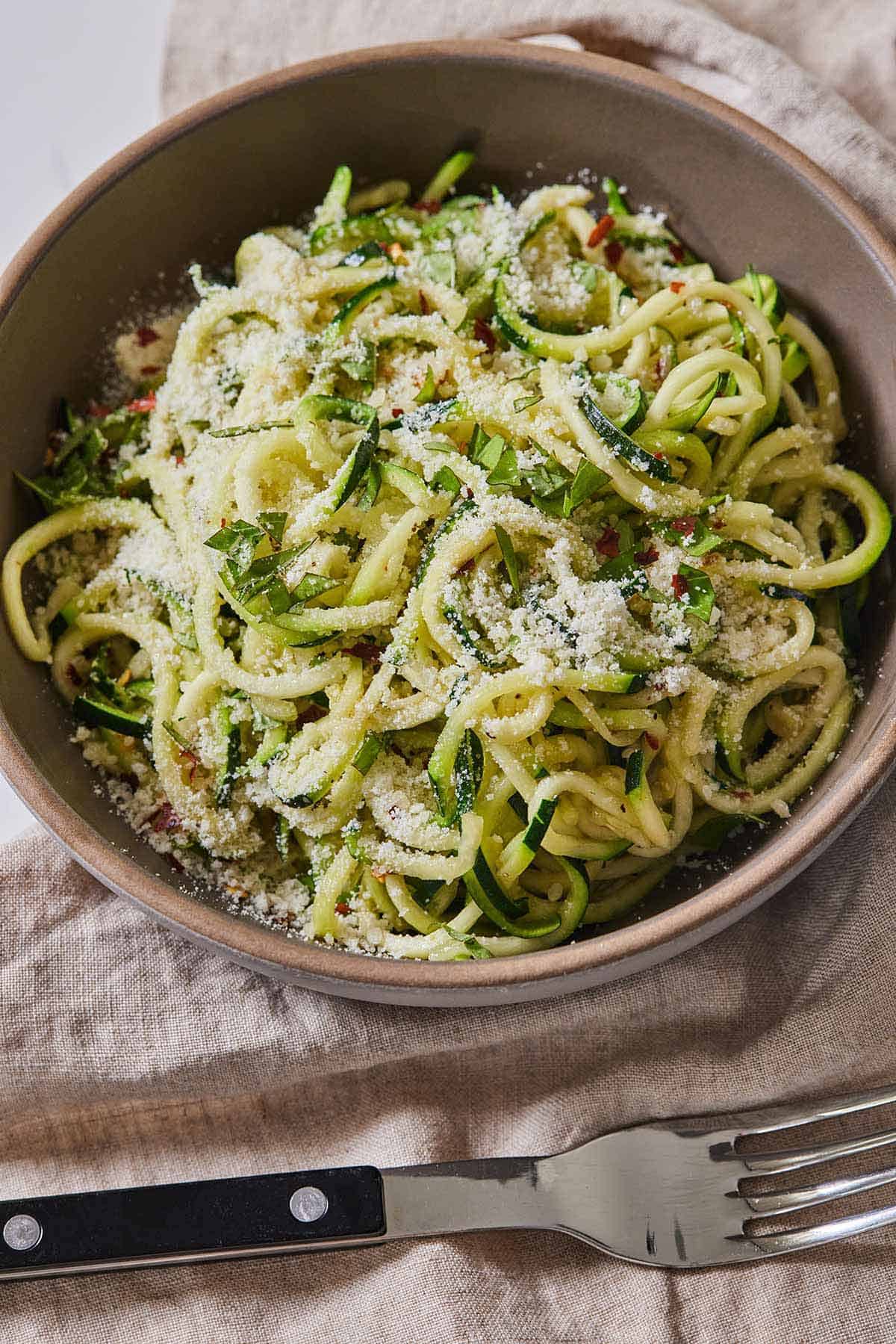 A bowl of zucchini noodles with parmesan and basil on top with a fork beside it.