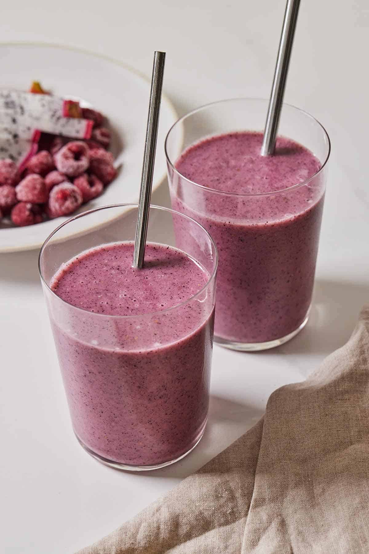 Two glasses of dragon fruit smoothies with fruit in the background.