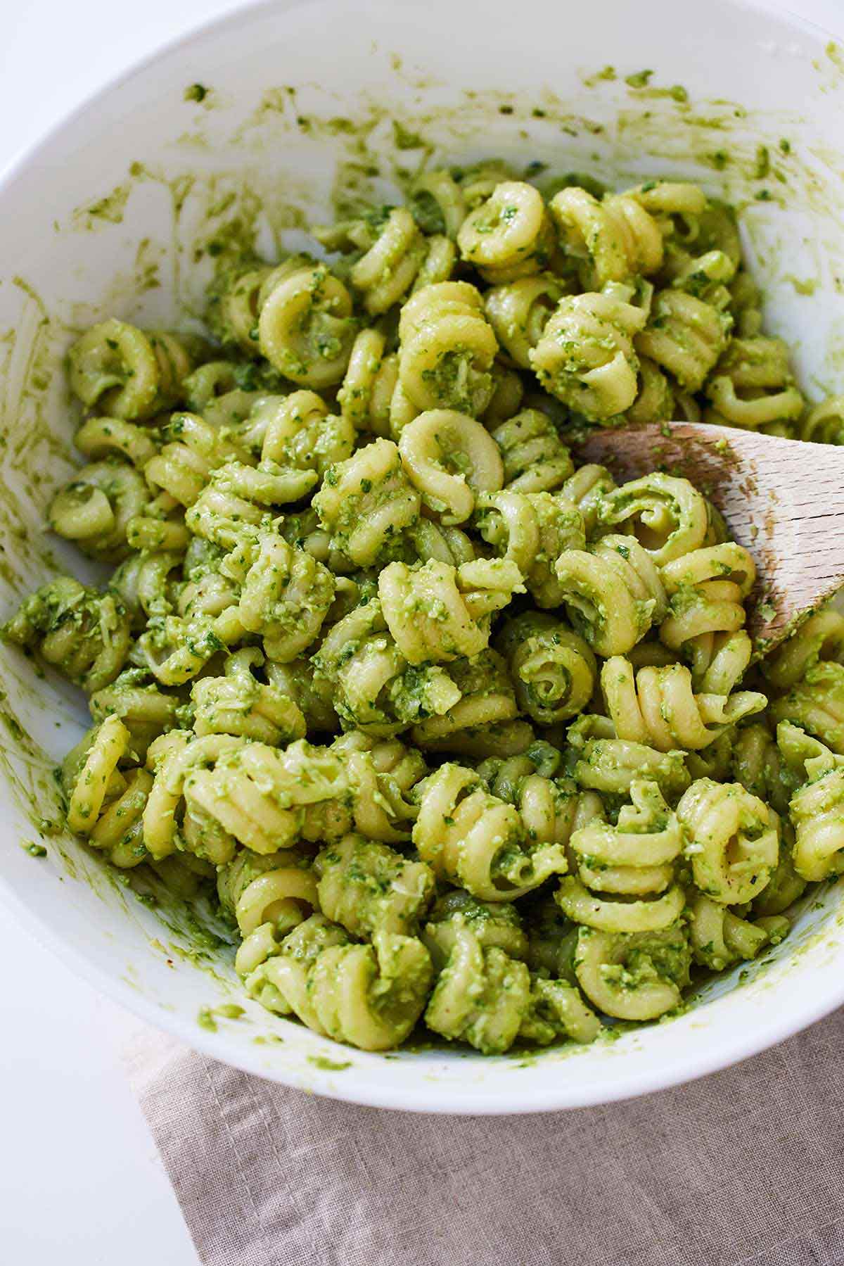 A bowl of avocado pesto pasta with a wooden spoon mixing it.