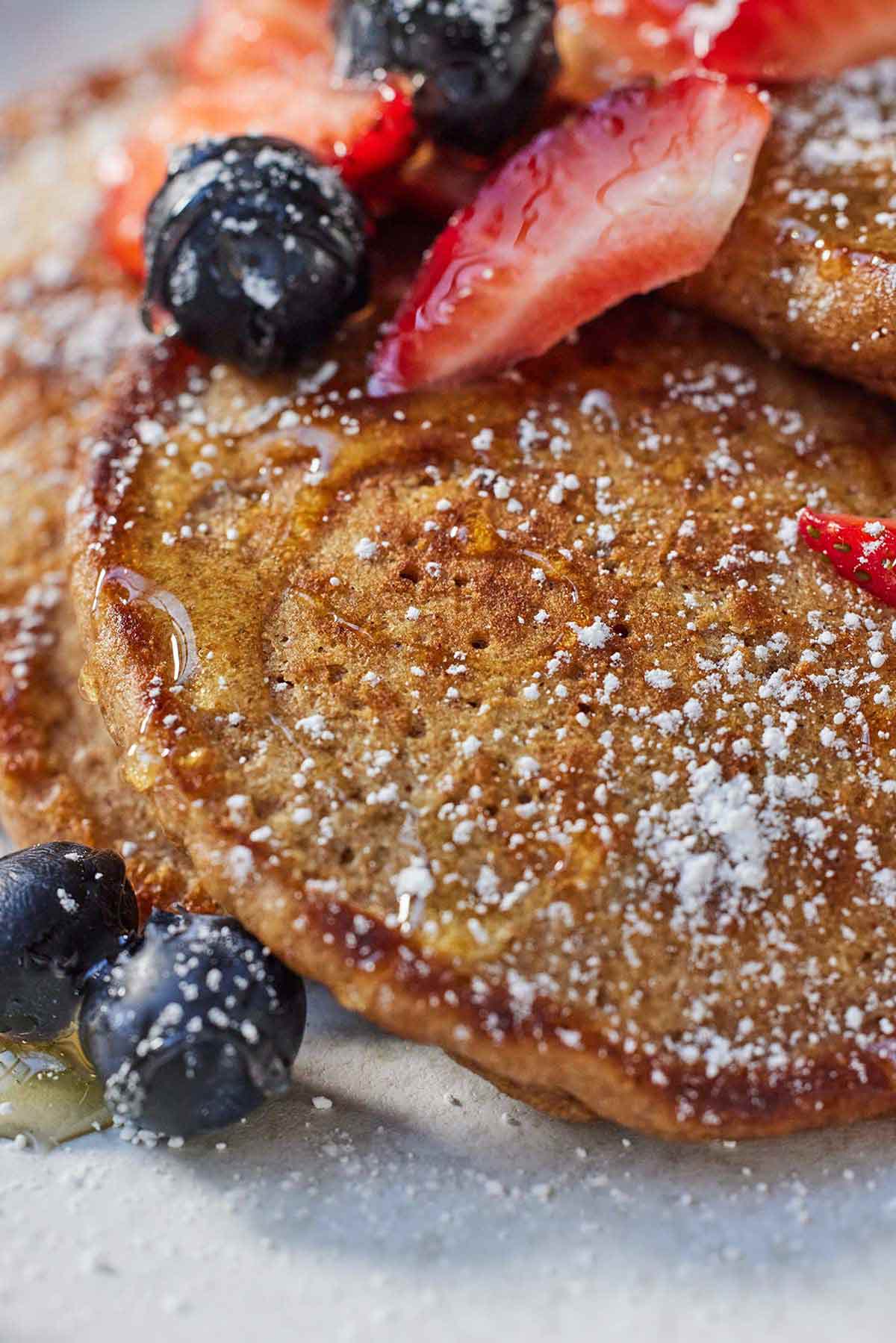 Close up of whole wheat pancakes with powdered sugar, syrup, and fruit.