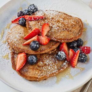 A plate of whole wheat pancakes with fruit, syrup, and powdered sugar.