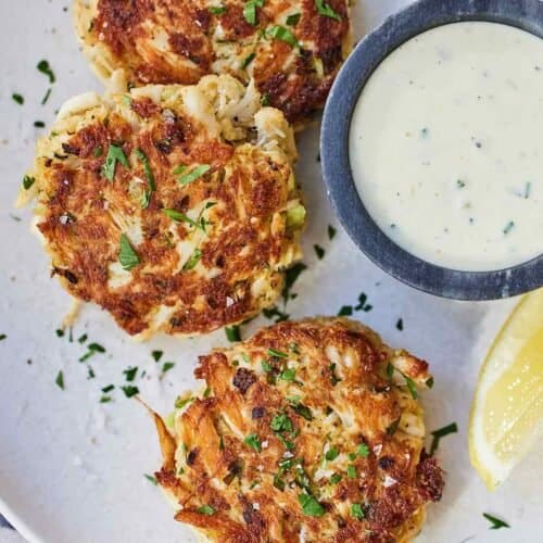 A plate with three crab cakes with a bowl of tartar sauce on the side.