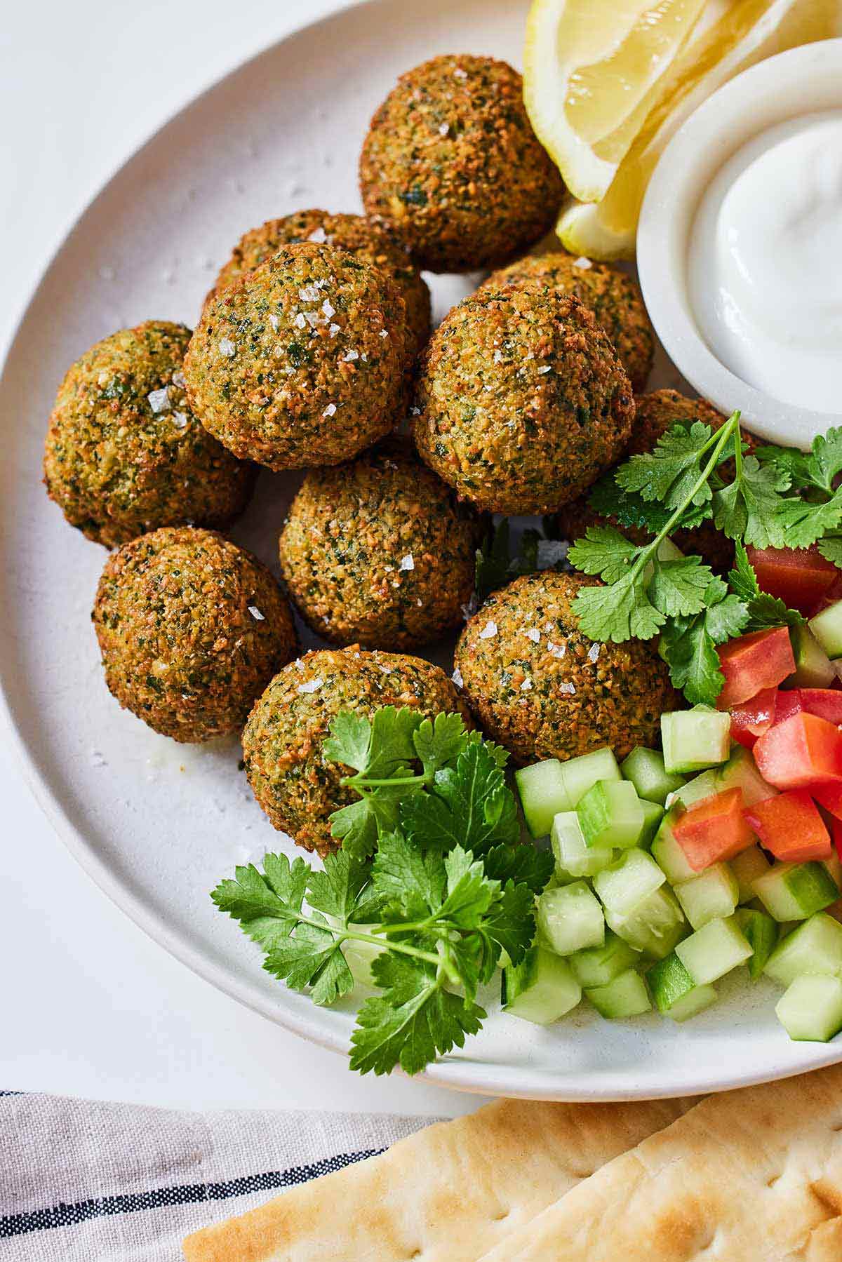 A plate with multiple falafels alongside fresh herbs, cucumbers, lemon wedges, and dip.