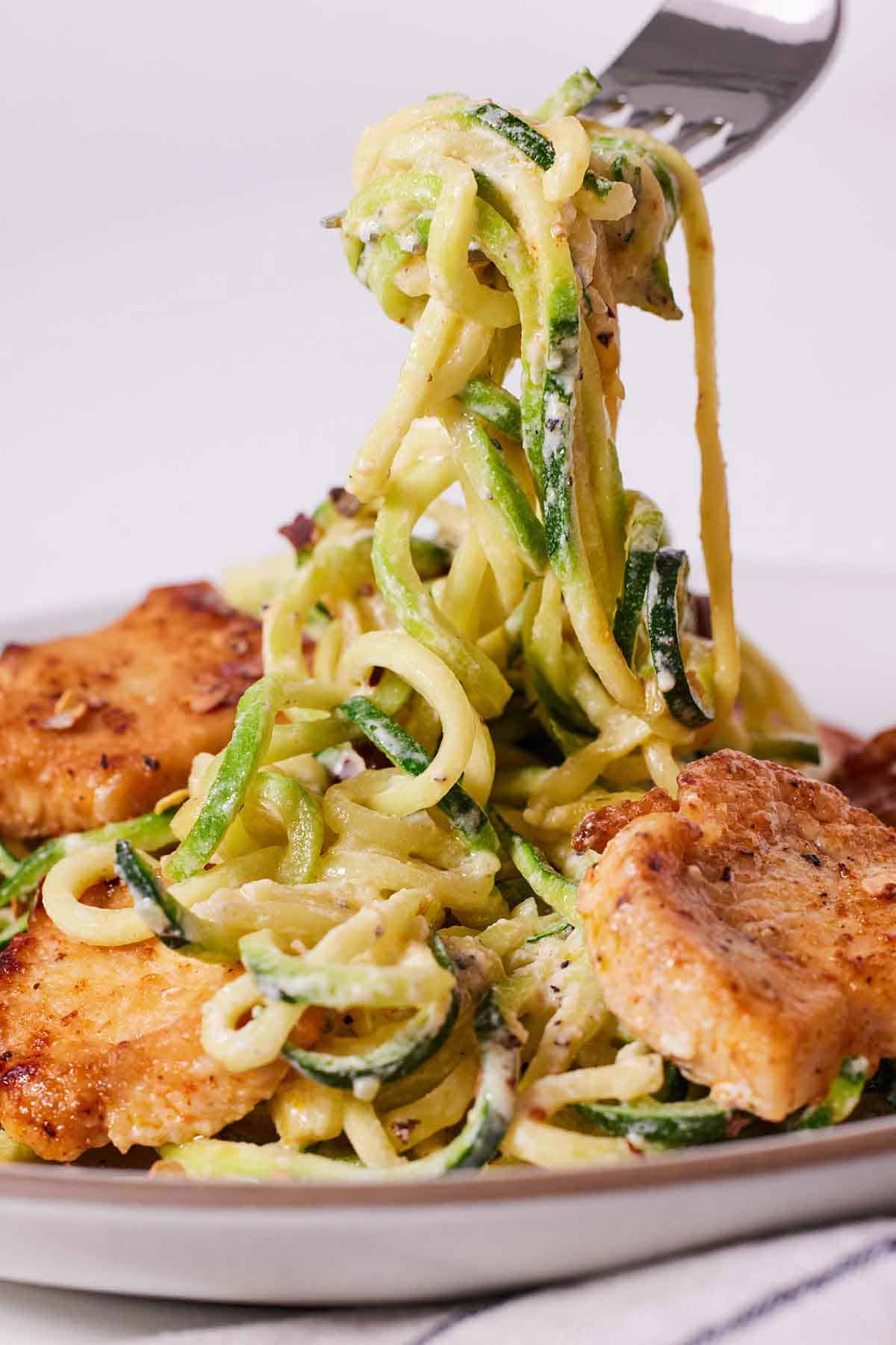 A forkful of zucchini noodles lifted from a plate of chicken Alfredo.