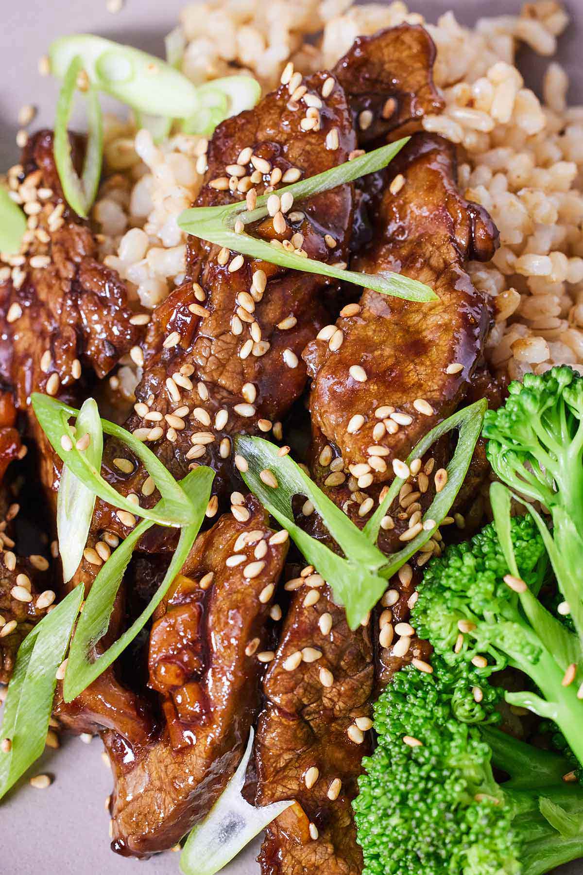 Close up view of orange beef with green onions and sesame seeds beside some broccoli and rice.