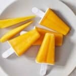 A plate with four mango popsicles with two slices of mango on the side.