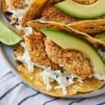 A plate with air fryer fish tacos with a focus on the crispy fish filling with sliced avocado and coleslaw.