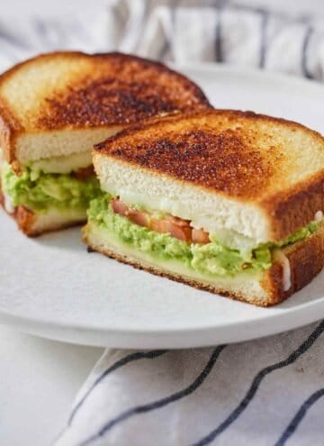 An avocado grilled cheese cut in half on a white plate showing the cheese, avocado, and tomato filling.