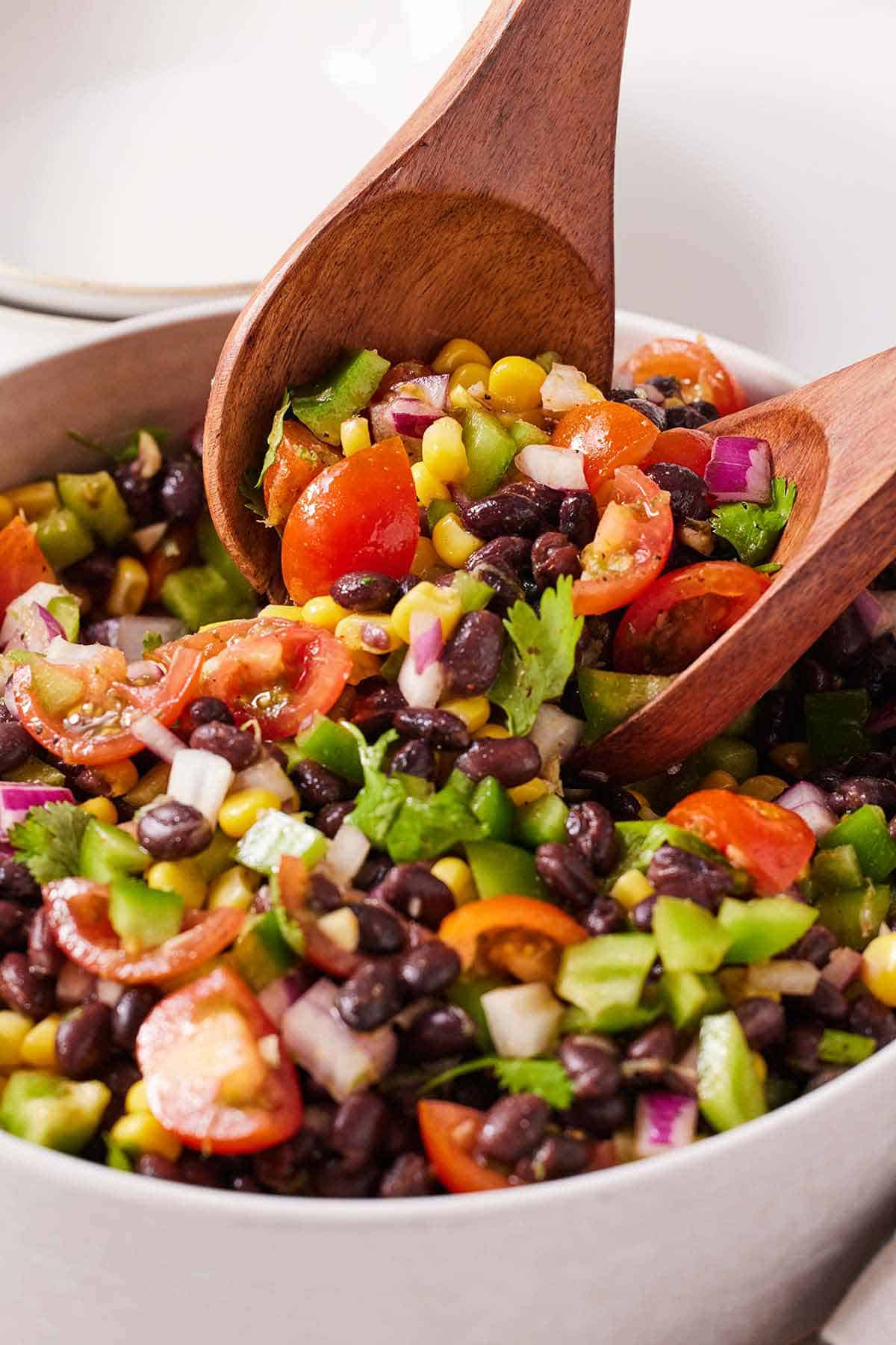 A bowl of black bean salad with two large wooden spoons scooping it up.