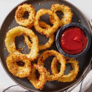 Overhead view of a dark plate with nine golden onion rings of assorted sizes by a bowl of ketchup.